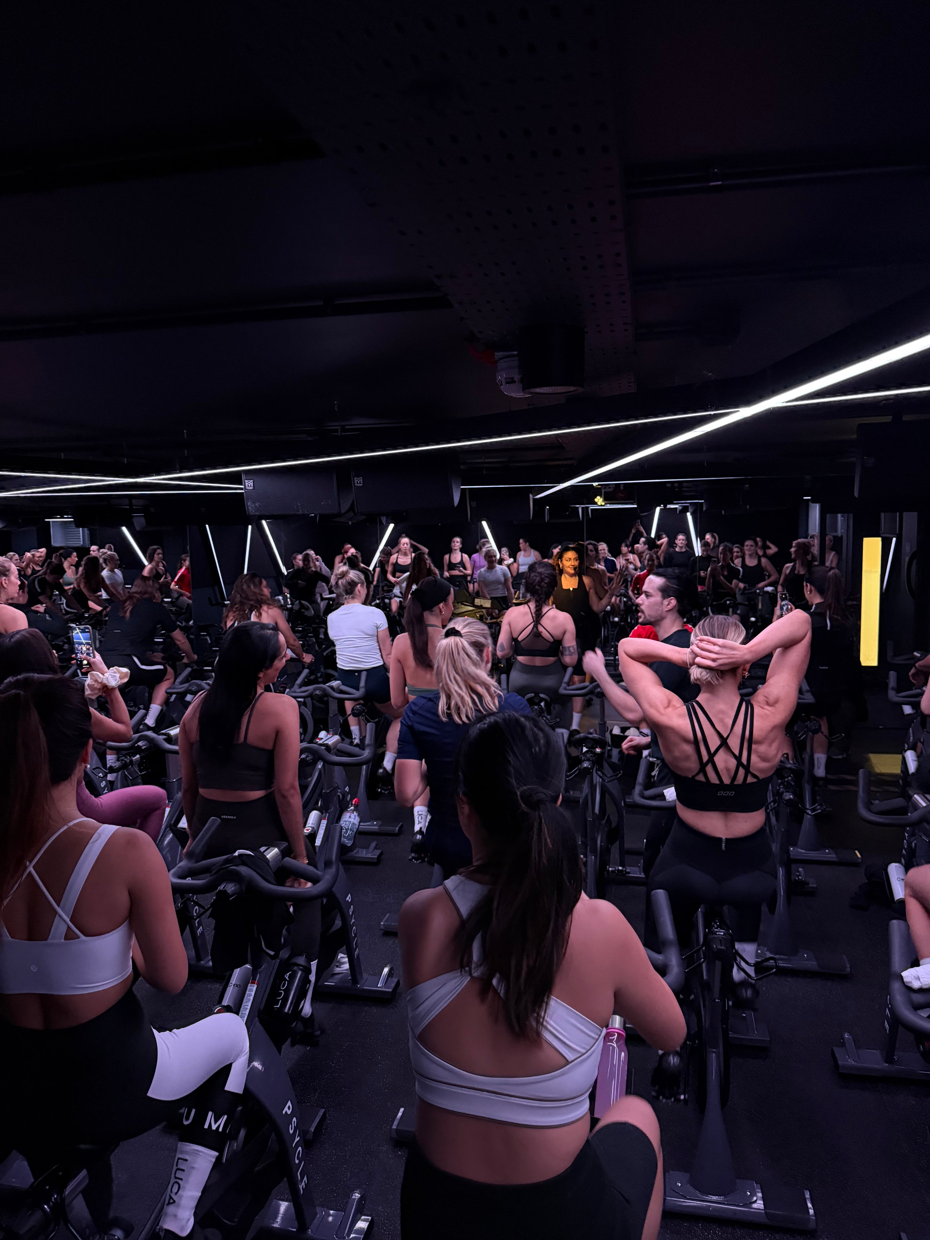 Packed-indoor-cycling-studio-during-a-class-viewed-from-behind-Dozens-of-participants-mostly-women-are-cycling-in-various-sports=bras-and-activewear-tops- including-cross-back-and-racerback-styles-The-studio-is-dark- with-overhead-white-neon-accent- lighting