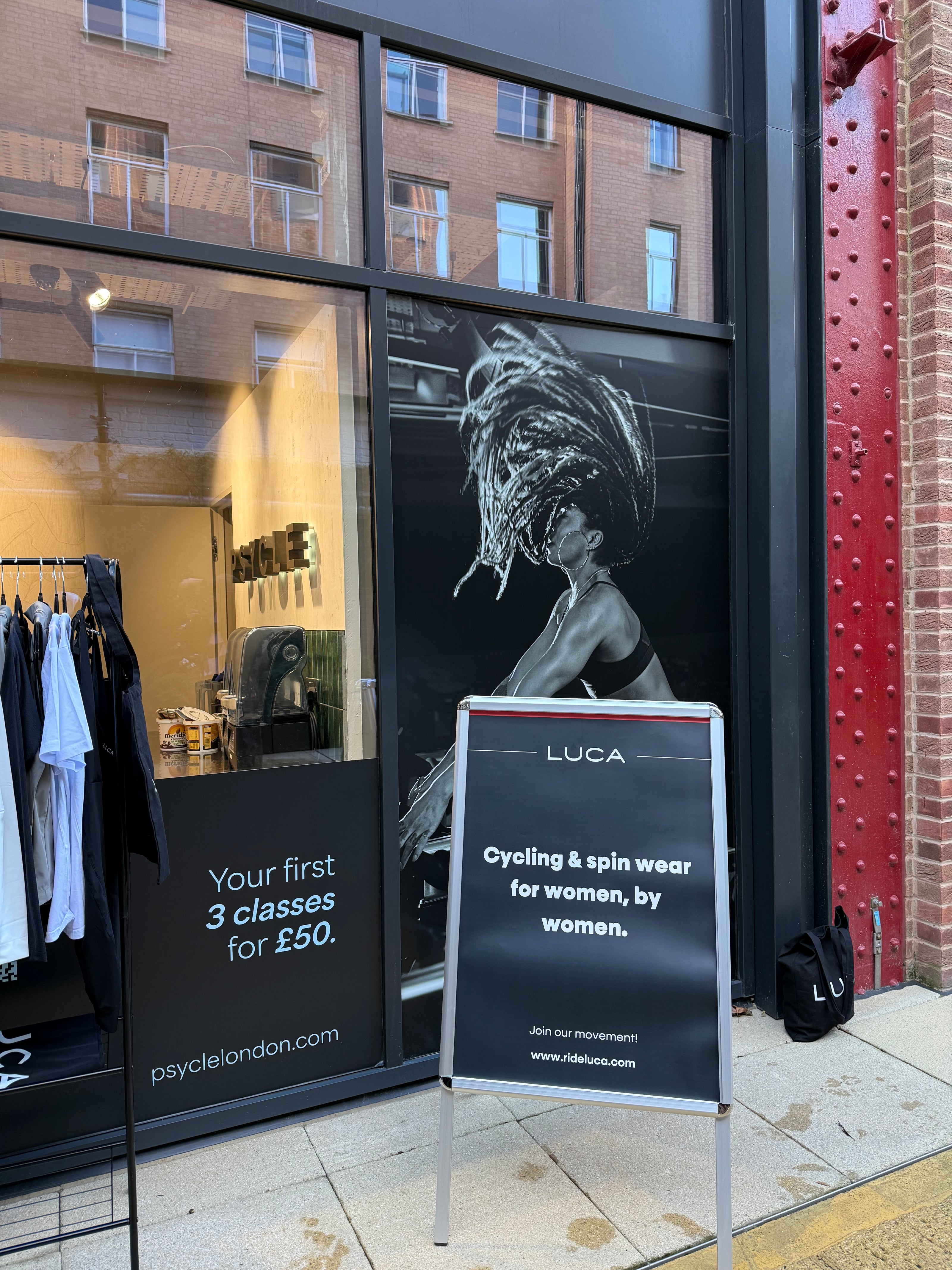 Storefront-window-featuring-a-large-black-and-white-image-of-a- woman-with-dynamic-hair-next-to- retail-clothing-racks-A-prominent-A-frame-sign-promotes-the-brand- 'LUCA'-with-the-text-"Cycling-&-spin-wear-for-women-by-women,"- and-a-website-address-The-window-also-advertises-a-promotion"-Your-firs-3-classes-for- £50