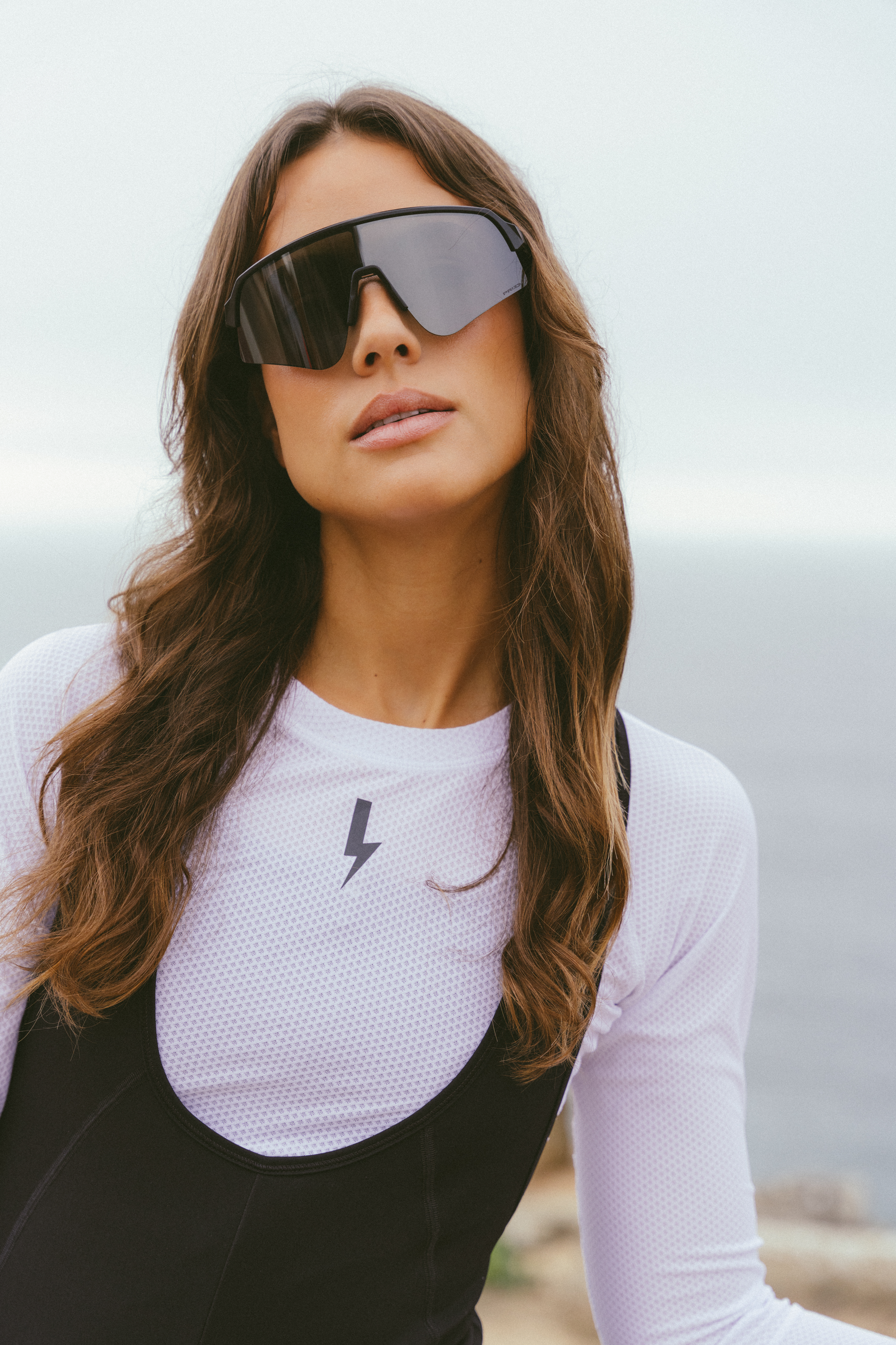 Close-up-of-a-woman-outdoors- wearing-large-black-wrap-around- cycling-sunglasses-with-a-single- mirrored-lens-She-is-wearing-a-white-long-sleeved-Radiance-base- layer-top-with-a-subtle-mesh-texture-and-a-small-black-lightning- bolt-sign-centered-on-the-chest- The-top-is-layered-beneath-black- bib-shorts-visible-at-the-bottom-The-background-is-a-bright-slightly- blurred-outdoor-scene-by-the-water