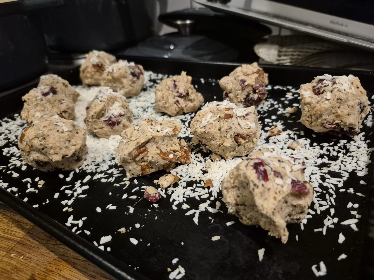 Unbaked-cookie-dough-balls- containing-visible-ingredients-like- pecans-dried-cranberries-and-poppy-seeds-dusted-with-shredded- coconut-on-a-black-baking-tray- ready-for-the-oven