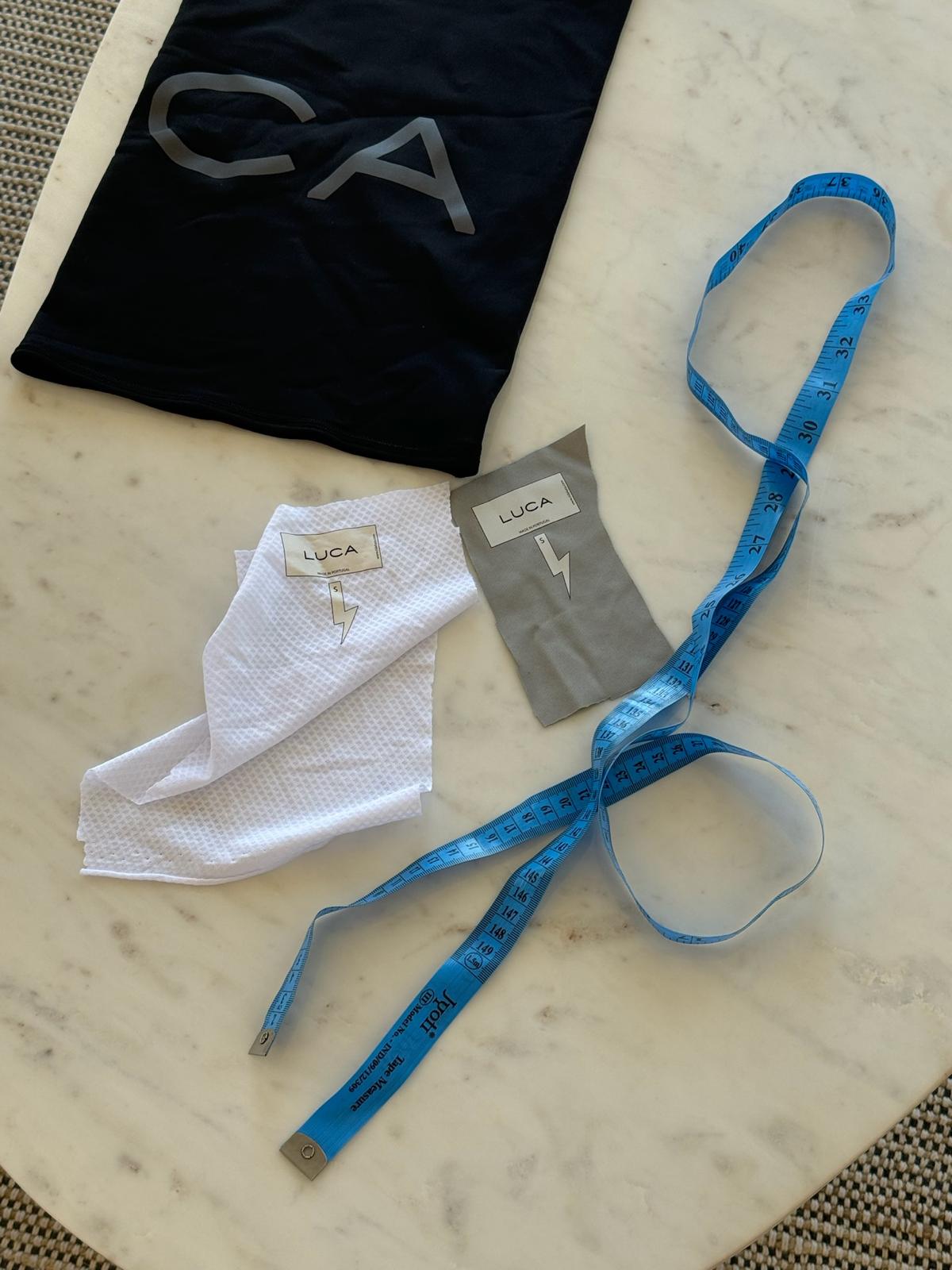 A-flat-lay-image-of-various-items- displayed-on-a-white-marble- surface-The-items-include-a-black- fabric-item-possibly-a-sleeve-with- the-silver-print-CA-visible-part-of- the-word-LUCA-There-is-also-a- folded-white-textured-fabric-swatch-and-a-small-gray-fabric- swatch-both-featuring-a-white-tag- with-the-brand-name-LUCA-and-a- lightning-bolt-icon-A-bright-blue- tape-measure-is-laid-diagonally- across-the-items