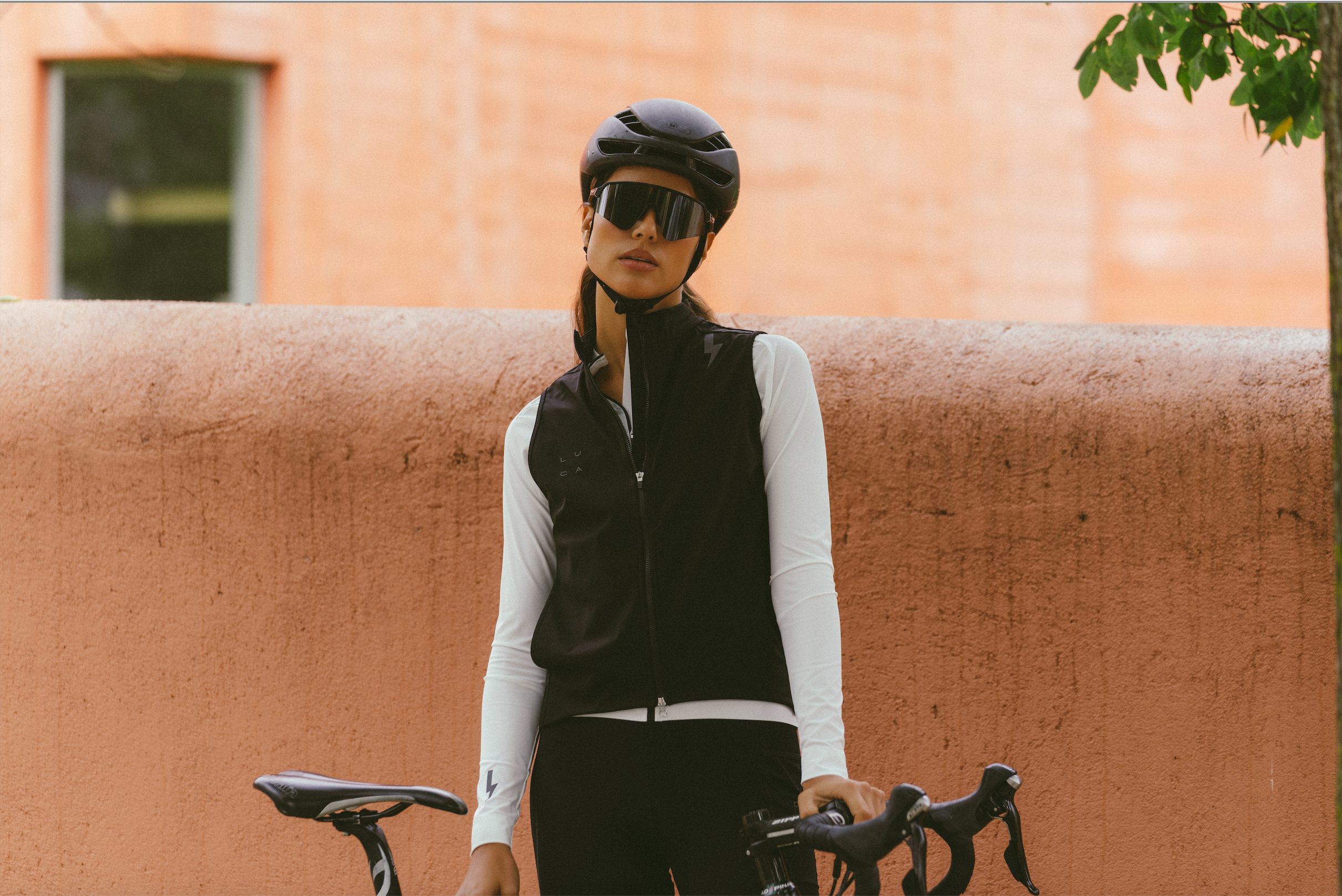 Model-wearing-a-black-cycling-vest- with-a-white-lightning-bolt-sign-over-a-white-long-sleeve-top-ready-to-ride-with-a-black-helmet- and-sunglasses