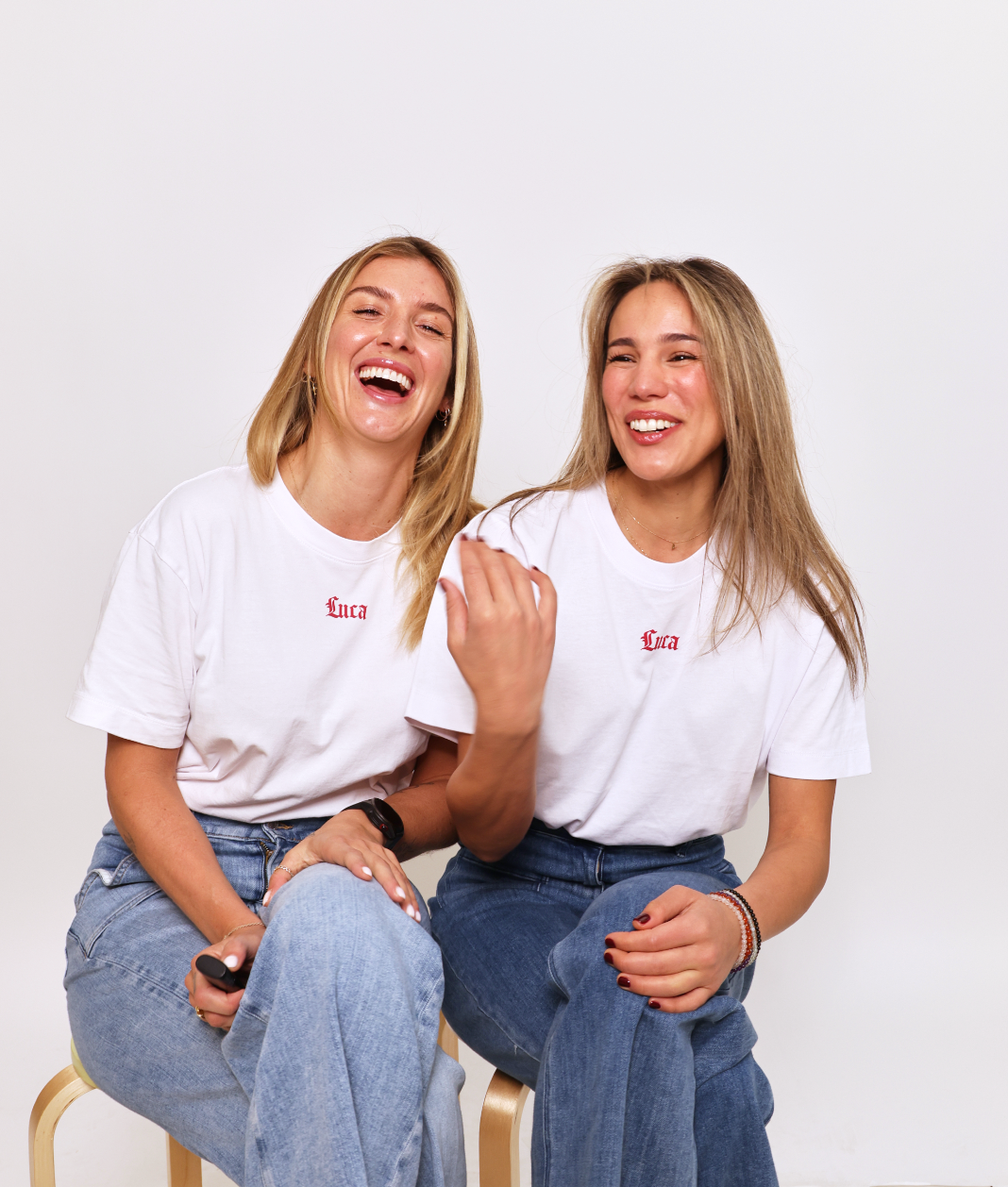 Two-smiling-women-sitting-together-and-laughing-in-a-studio- shot-They-are-both-wearing- matching-white-crewneck-t-shirts- that-feature-the-word-"Luca"-printed-in-small-red-script-on-the- chest-paired-with-denim-jeans