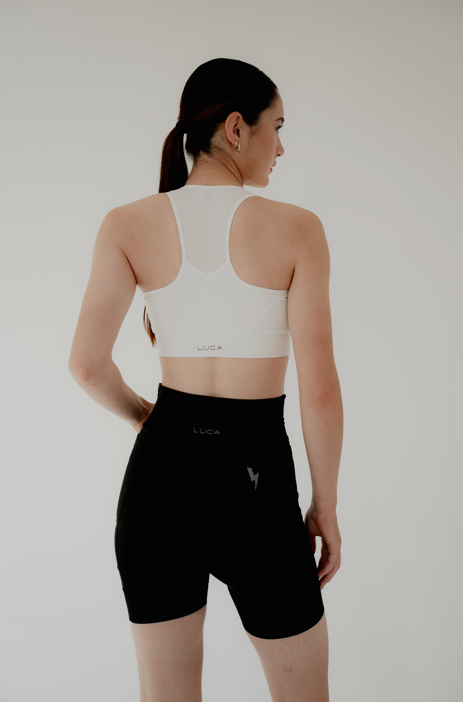 Back-view-of-a-woman-modeling-a-white-spots-bra-with-mesh-panel-and-LUCA-logo-paired-printed-with-black-high-waisted-biker-shorts-featuring-a-reflective-lighting-bolt-graphic