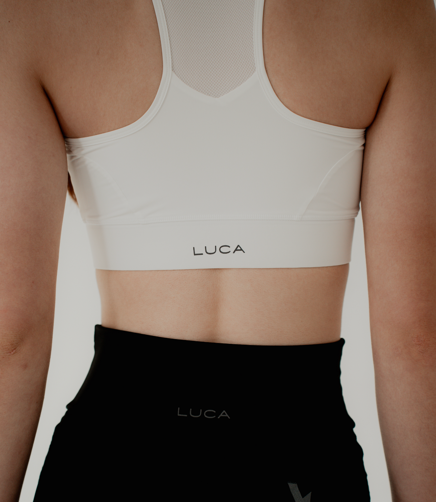 A-close-up-back-view-shot- highlighting-the-design-details-of- the-off-white-sports-bra-and-black- athletic-shorts-The-sports-bra- features-a-racerback-design-with-a- sheer-off-white-mesh-panel-at-the-top- The-"LUCA"-brand-logo-is-centered-in-black-on-both-the- bottom-band-of-the-bra-and-the- shorts-emphasizing-the-matching- activewear-set