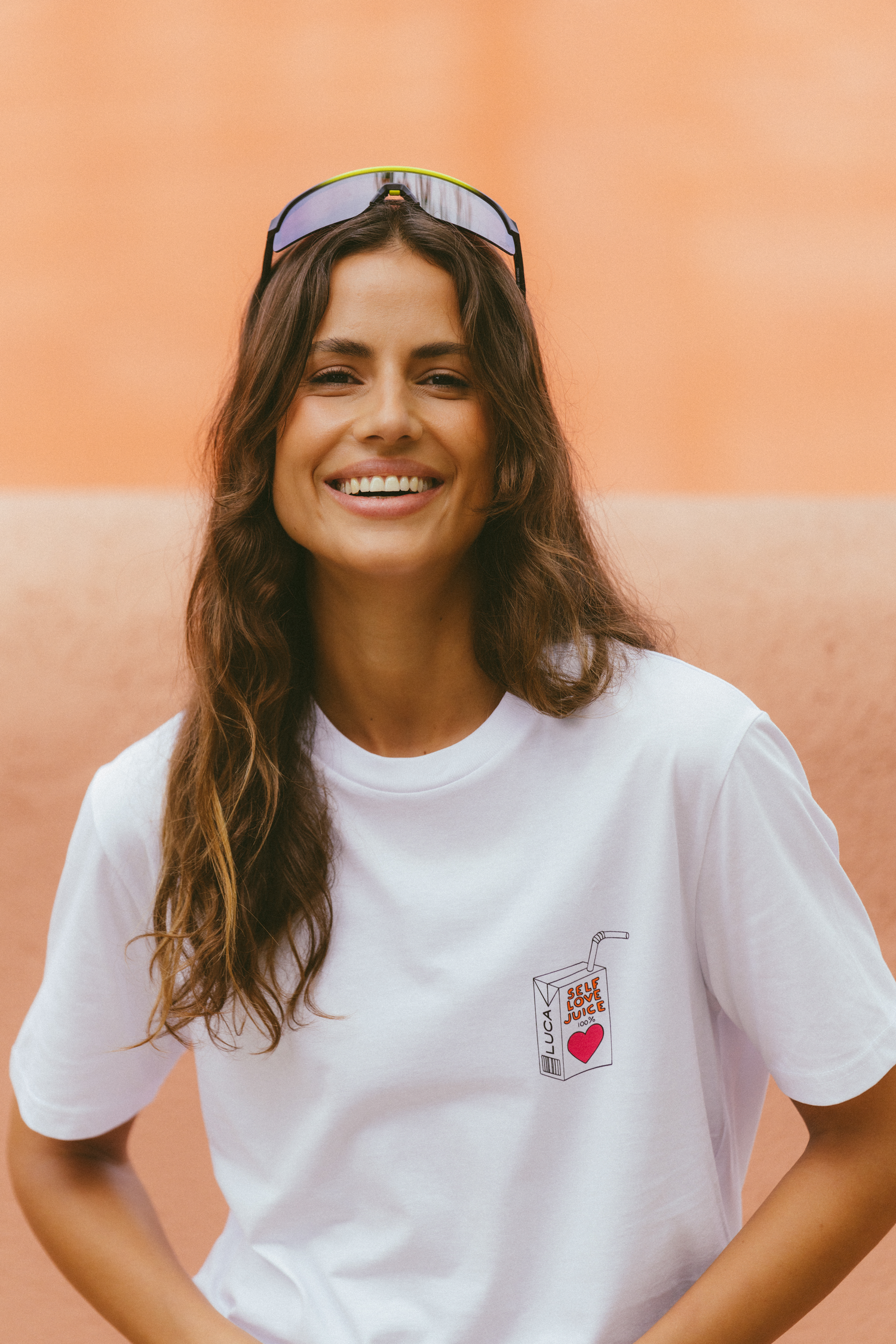 A-smiling-woman-with-long-brown- hair-wearing-a-white-crewneck-t-shirt-The-shirt-features-a-small- embroidered-design-of-a-juice-box- on-the-chest-labeled-SELF-LOVE- JUICE-100%-with-a-red-heart-She-is-wearing-cycling-sunglasses- pushed-up-on-her-head-and-is- standing-against-a-terracotta-colored-background