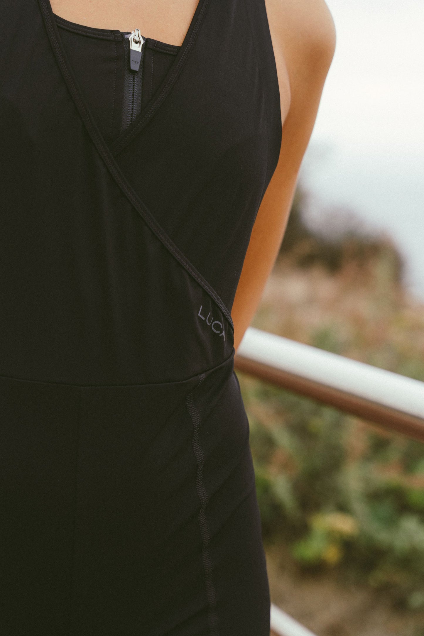 Extreme-close-up-shot-of-the-torso- area-of-a-black-athletic-skinsuit- trisuit-The-garment-features-a-black-on-black-embroidered-logo- reading-"LUCA"-near-the-waist-hip- below-a-cross-over-neckline-with-a- visible-silver-zipper-The-background-is-blurred-outdoors-with-foliage-and-a-railing