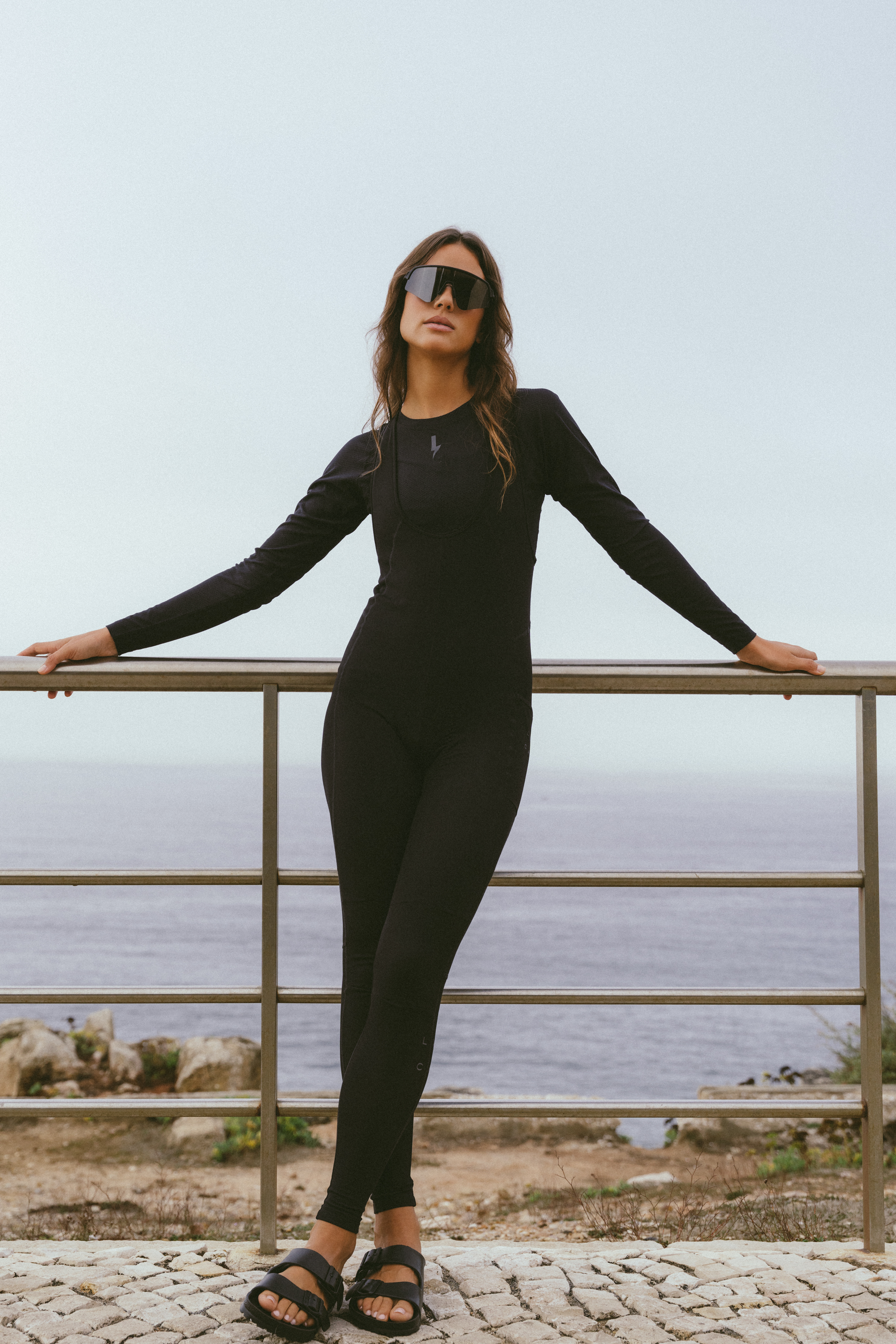Full-length-shot-of-a-woman- wearing-a-black-long-sleeved- Stride-Bib-Tightwith-a-subtle-ribbed-texture-The-garment-is- accessorized-with-large-dark-wrap-around-athletic-sunglasses-and- black-two-strap-sandals-A-small- black-lightning-bolt-logo-is-visible- near-the-neckline-She-is-leaning- against-a-metal-railing-overlooking- the-ocean-on-a-cloudy-day- standing-on-cobblestone-pavement