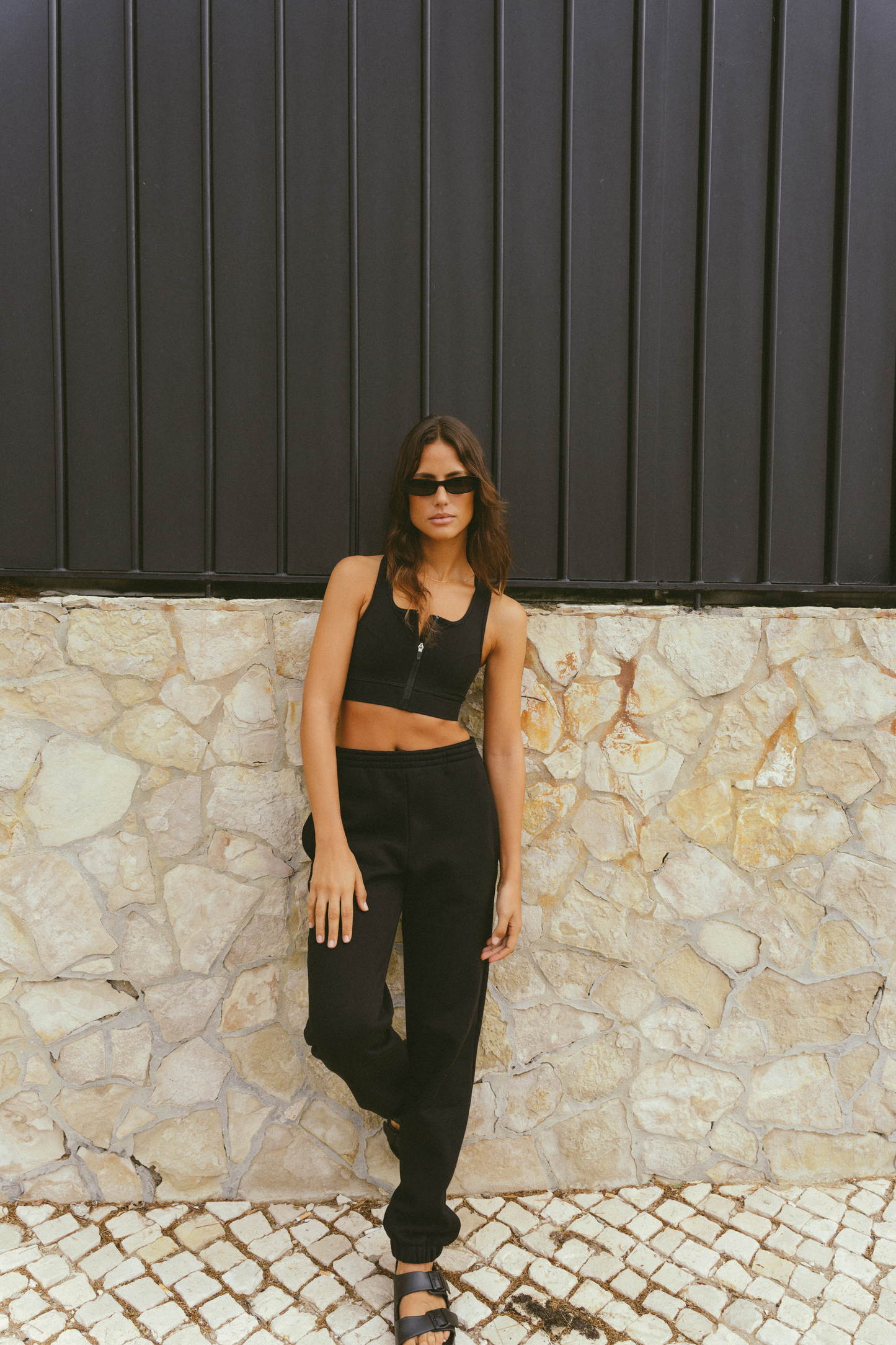 Woman-wearing-a-black-cropped-tank-top-and-black-jogger-pants-standing-against-a-stone-wall-with-one-leg-slightly-bent-and-wearing-black-sandals