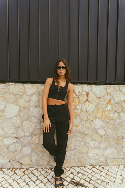 Woman-wearing-a-black-cropped-tank-top-and-black-jogger-pants-standing-against-a-stone-wall-with-one-leg-slightly-bent-and-wearing-black-sandals