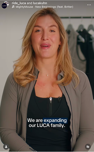 Close-up-of-a-woman-with-long- blonde-hair-looking-directly-at-the- camera-She-is-wearing-a-high-collared-taupe-dark-gray-athletic- jacket-open-at-the-neck-layered- over-a-black-zip-front-technical-top- A-rack-of-dark-clothing-is-visible-in- the-blurred-background