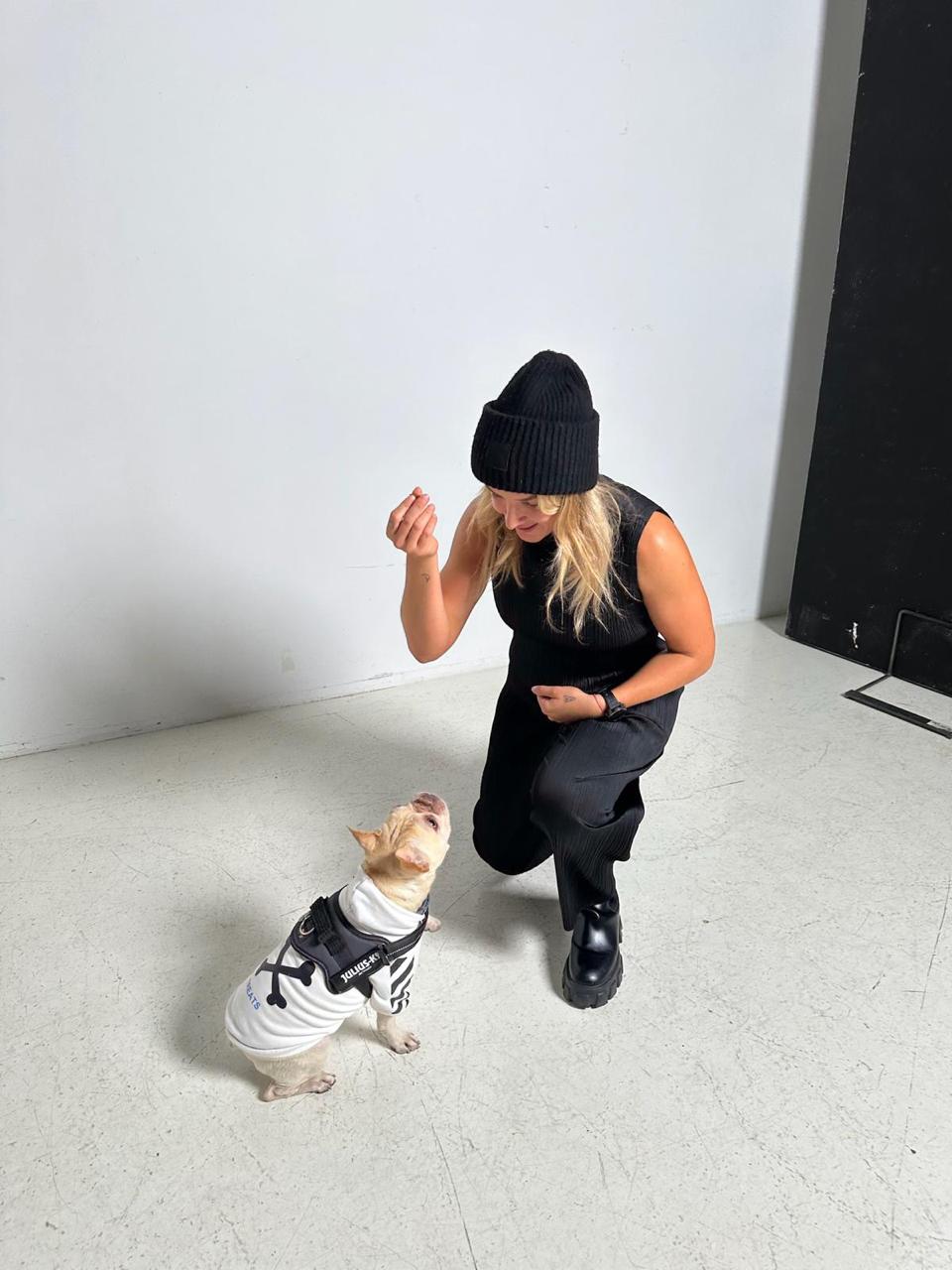 Woman-in-all-black-clothing-kneeling-to-interact-with-a-small- dog-wearing-a-white-striped-outfit- and-harness-in-a-studio