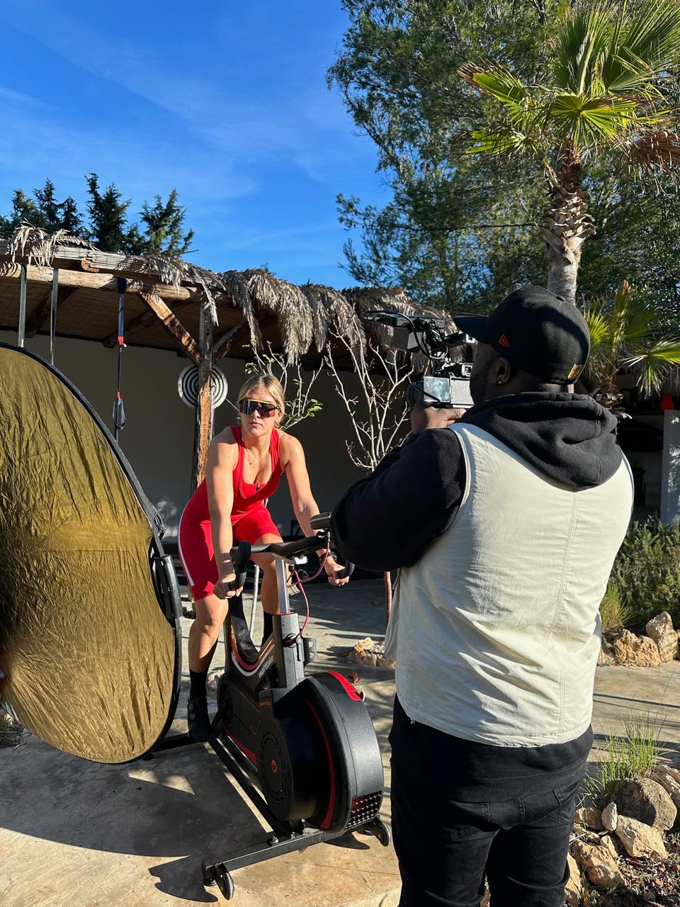 Woman-in-red-workout-gear-cycling-outdoors-while-being-filmed-by-a-camera-operator
