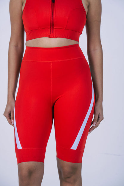 A-close-up-studio-shot-focusing-on- the-midsection-and-legs-of-a- woman-wearing-bright-red-high-waisted-cycling-shorts-The-shorts- feature-a-white-graphic-stripe-that- curves-diagonally-up-from-the-hem -on-both-outer-sides-of-the-legs- The-model-is-also-wearing-a- matching-red-sports-bra-crop-top- with-a-visible-front-zipper