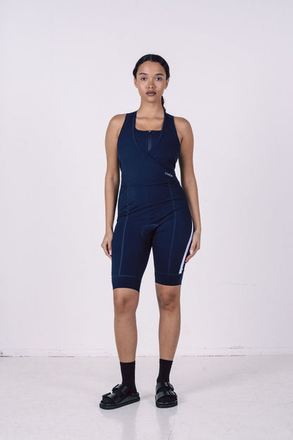 Woman-wearing-a-navy-blue-cycling-Road-All-In-One-with-a-zip-front-wrap-style-top-and-white- vertical-stripe-on-the-leg-Padded- one-piece-for-competitive-biking- and-training