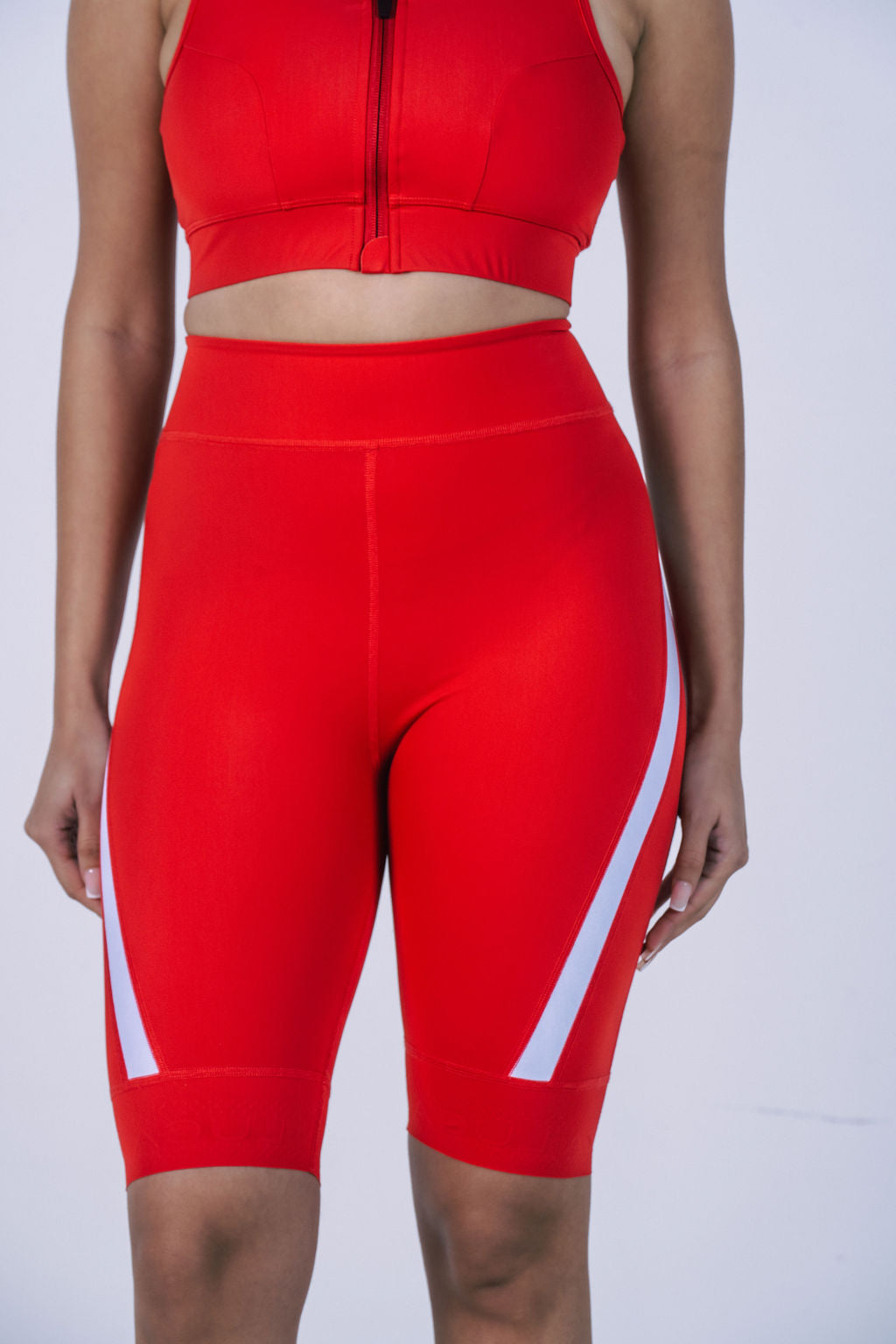 Close-up-of-a-woman-wearing-high-waisted-bright-red-biker-shorts-with-white-angled-stripe- accents-on-the-leg-paired-with-a- matching-zip-front-sports-bra-Mid-length-cycling
