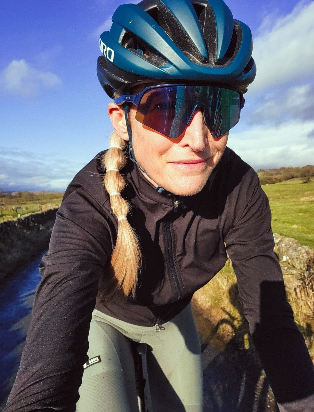 Close-up-photo-of-a-female-cyclist- wearing-a-teal-Giro-helmet-large- reflective-cycling-sunglasses-and-a- black-long-sleeved-cycling-jacket- She-is-smiling-slightly-while-riding-a- bike-on-a-rural-path-with-a stone- wall-and-green-fields-visible