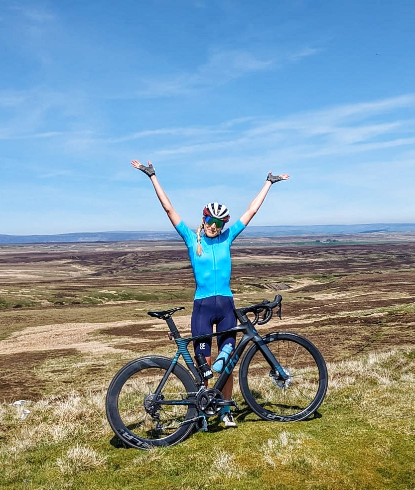 Woman-in-a-high-performance-blue-and-navy-cycling-kit- celebrating-a-successful-climb-next-to-a-modern-black-road-bike- with-deep-dish-wheels-and-blue- accents