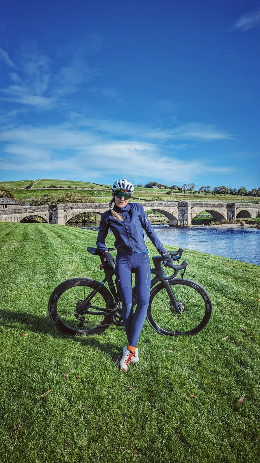 A-female-cyclist-in-a-matching-navy-blue-cycling-kit-standing-on-a- lush-green-lawn-next-to-her-black- road-bike-She-is-smiling-wearing-a- helmet-and-sunglasses-with-a- historic-stone-arch-bridge-spanning-a-river-visible-in-the-sunny-background