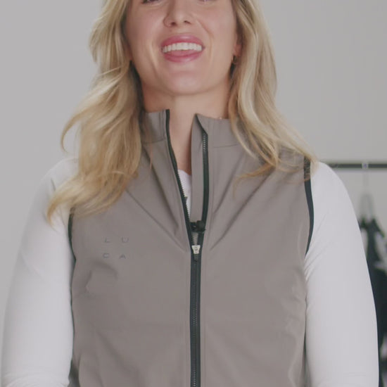 Video-demonstrating-the-features-of-the-Luca-Ascend-cycling-gilet-in-taupe-color-A-women-details-the-gilet-three-layer-windproof-and-water-repellemt-faric-moisture-wicking-inner-layer-doule-ended-zipper-silicone-gripper-hem-reflective-logos-and-the-ergonomic-cut-for-riding-comfort