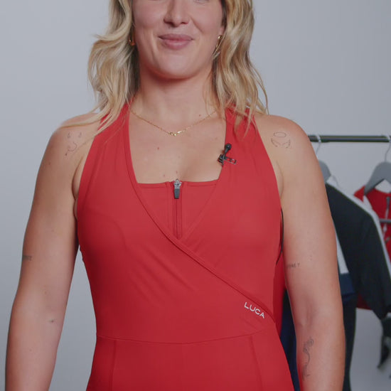 A-video-featuring-a-blonde-woman- presenting-and-discussing-the-features-of-a-red-cycling-road-all-in-one-The-garment-is-sleeveless-with-a- square-neckline-a-short-zipper-and-a- unique-crossover-front-section-for- coverage-and-support-The-back-panel-shows-a-keyhole-cutout-for- ventilation-and-white-reflective-stripes-on-the-straps-and-center-for- visibility-The-presenter-highlights-the women's-specific-chamois-anti-chafing-seams-silicone-leg-grips-and-the- use-of-high-strength-Italian-fabric