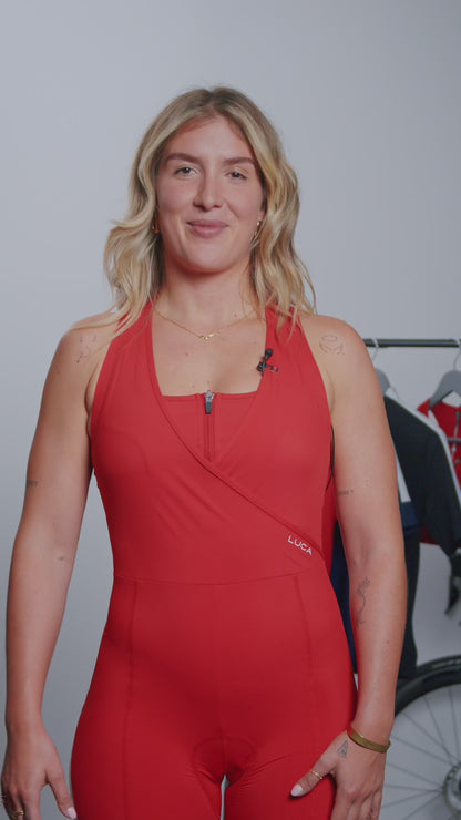 A-video-featuring-a-blonde-woman- presenting-and-discussing-the-features-of-a-red-cycling-road-all-in-one-The-garment-is-sleeveless-with-a- square-neckline-a-short-zipper-and-a- unique-crossover-front-section-for- coverage-and-support-The-back-panel-shows-a-keyhole-cutout-for- ventilation-and-white-reflective-stripes-on-the-straps-and-center-for- visibility-The-presenter-highlights-the women's-specific-chamois-anti-chafing-seams-silicone-leg-grips-and-the- use-of-high-strength-Italian-fabric