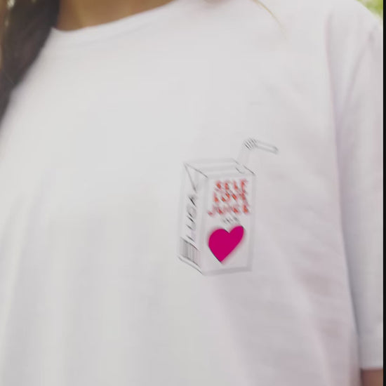 A-short-charming-video-clip-of-a-model-wearing-a-white-crew-neck-t-shirt-with-a-unique-grahic-print-The-print-located-on-the-left-chest-depicts-a-stylized-juice-box-that-reads-SELF-LOVE-JUICE-with-a-pink-heart-graphic-and-the-brand-name-LUCA-The-model-smiles-and-blows-a-kiss-toward-the-camera-in-a-vibrant-outdoor-setting-highlighting-the-casual-fit-and-positive-message-of-the-comfortable-cotton-tee