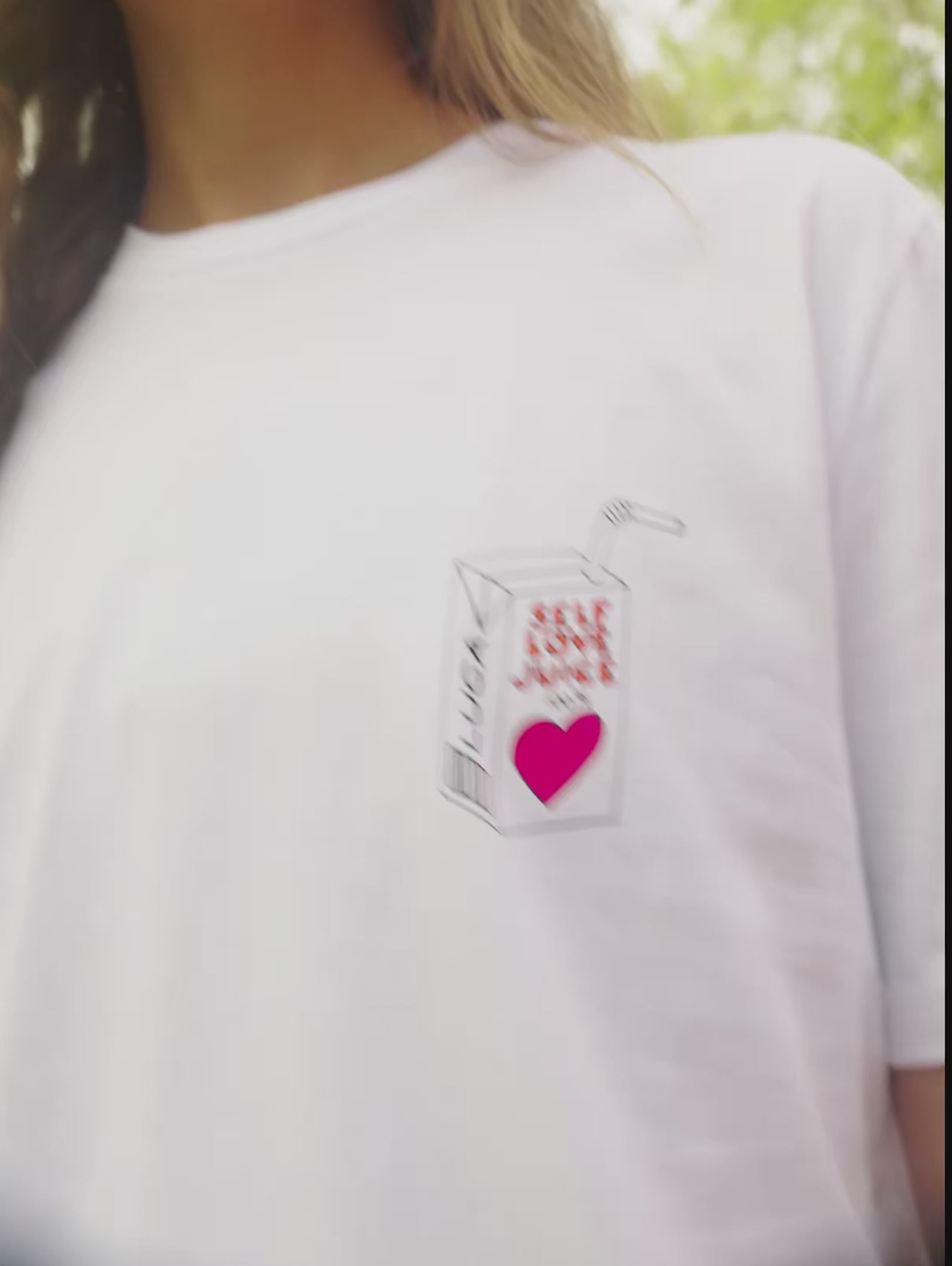 A-short-charming-video-clip-of-a-model-wearing-a-white-crew-neck-t-shirt-with-a-unique-grahic-print-The-print-located-on-the-left-chest-depicts-a-stylized-juice-box-that-reads-SELF-LOVE-JUICE-with-a-pink-heart-graphic-and-the-brand-name-LUCA-The-model-smiles-and-blows-a-kiss-toward-the-camera-in-a-vibrant-outdoor-setting-highlighting-the-casual-fit-and-positive-message-of-the-comfortable-cotton-tee